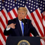 A man in a suit speaking at a podium with American flags in the background