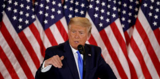 A man in a suit speaking at a podium with American flags in the background