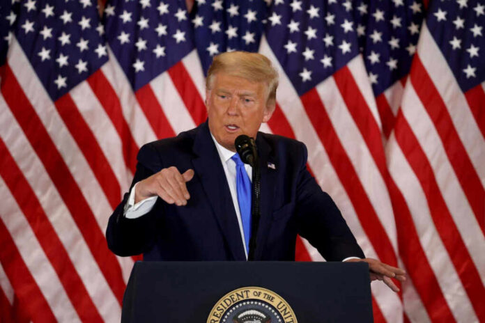 A man in a suit speaking at a podium with American flags in the background