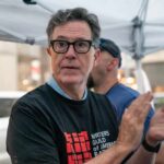 A man in a black t-shirt with a writers' guild logo clapping at a public event