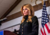 A woman in formal attire speaking at a podium with an American flag in the background