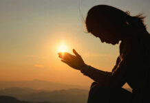 Silhouette of a person in a meditative pose with hands reaching towards the sun during sunset
