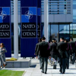 Military personnel walking past NATO banners with two women conversing in the foreground