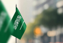 A green flag of Saudi Arabia with white Arabic text and a sword, set against a blurred background