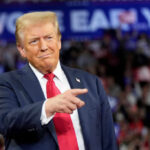 A man in a suit gestures while speaking at a political rally with a cheering crowd in the background