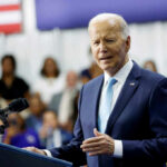 A man in a suit speaking at a podium with a microphone, an audience in the background