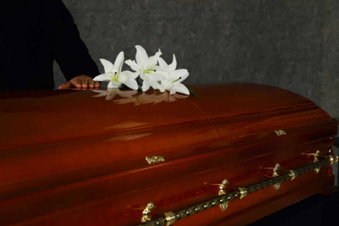 A wooden casket adorned with white lilies, with a hand resting on it