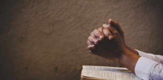 Hands clasped in prayer over an open book on a wooden table