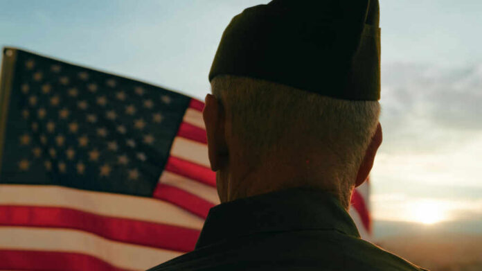 shutterstock_2513309275.jpg A veteran in uniform standing in front of an American flag during sunset