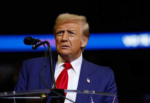 Trump’s Charlottesville Return SPARKS FURY A man in a blue suit and red tie speaking into a microphone on stage