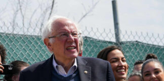 A group of people at an outdoor political rally, with a smiling man speaking and a woman laughing in the foreground