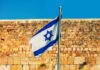 Israeli flag waving in front of a stone wall under a clear blue sky
