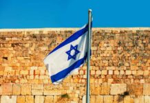 Israeli flag waving in front of a stone wall under a clear blue sky