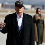 A political figure in a black coat and MAGA hat gesturing outdoors with a supporter in the background