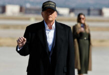 A political figure in a black coat and MAGA hat gesturing outdoors with a supporter in the background