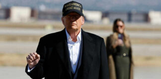 A political figure in a black coat and MAGA hat gesturing outdoors with a supporter in the background