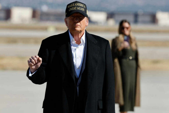 A political figure in a black coat and MAGA hat gesturing outdoors with a supporter in the background
