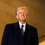 A man in formal attire standing confidently indoors with a serious expression