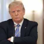 Shocking Podcast: Trump’s Life Allegedly Threatened A serious-looking man in a suit with arms crossed during a meeting