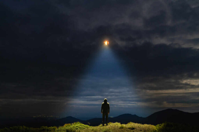 A mysterious figure standing on a mountain under a dark sky with a beam of light from a UFO