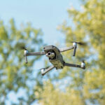A drone flying against a backdrop of green trees and a blue sky