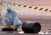 Hazmat worker kneeling near a fallen barrel at a cordoned-off site