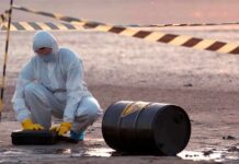 Hazmat worker kneeling near a fallen barrel at a cordoned-off site