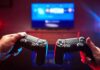 Two hands holding game controllers in front of a TV screen
