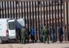 Border agents escort a group near a tall fence and a patrol van