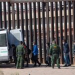 Border agents escort a group near a tall fence and a patrol van