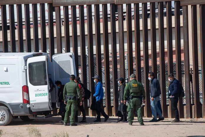 Border agents escort a group near a tall fence and a patrol van