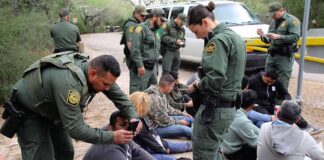 Law enforcement officers process a seated group outdoors