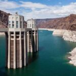 A large dam structure with two towers over a blue water reservoir surrounded by rocky mountains