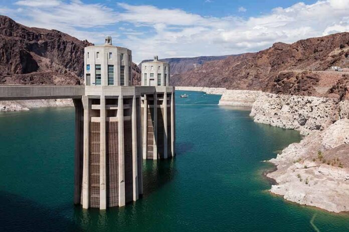 A large dam structure with two towers over a blue water reservoir surrounded by rocky mountains