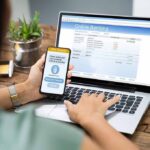 Person using smartphone to approve bank transfer beside laptop with online banking