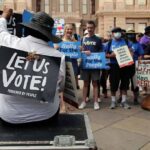Christian Jones sits with a 'Let Us Vote' sign facing a crowd at a voting rights rally