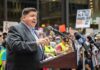 Federal Showdown Erupts Over Chicago Speaker at a podium addressing a crowd with microphones and protest signs