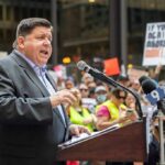 Federal Showdown Erupts Over Chicago Speaker at a podium addressing a crowd with microphones and protest signs
