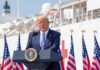 Donald Trump speaking at a podium with American flags and a ship in the background