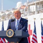 Donald Trump speaking at a podium with American flags and a ship in the background