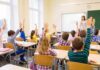 Students raise hands in a classroom while a teacher stands at the front