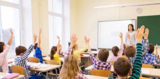Students raise hands in a classroom while a teacher stands at the front