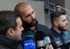 Man speaking to reporters with microphones outside a building