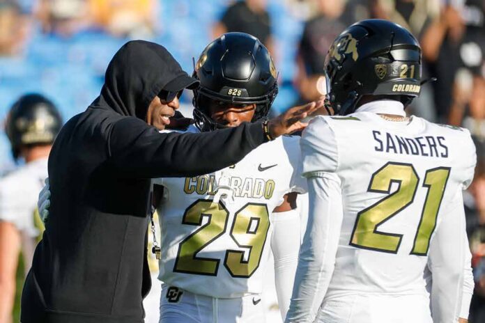 Coach in black hoodie speaks to two football players in white uniforms